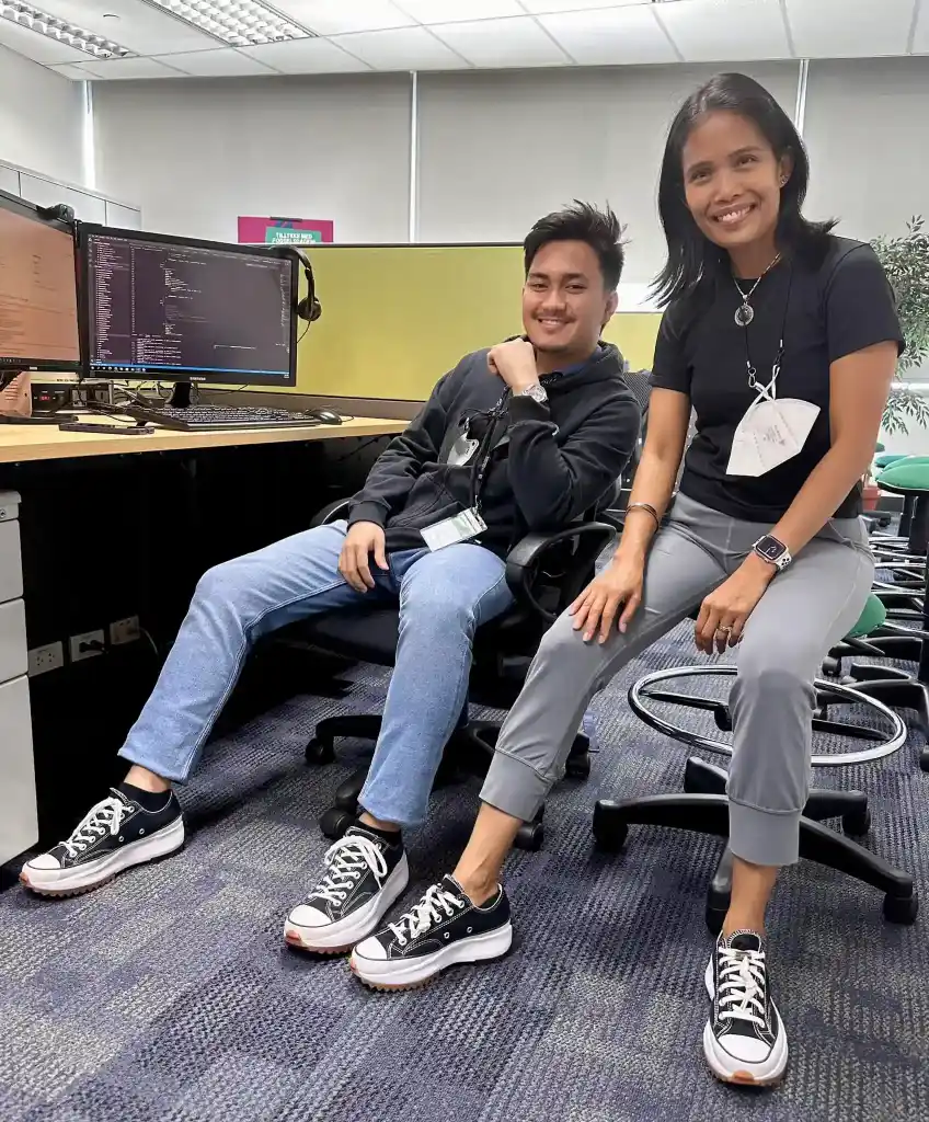 Two colleagues in an office wearing matching black and white platform Chuck Taylor sneakers, seated near computer monitors.