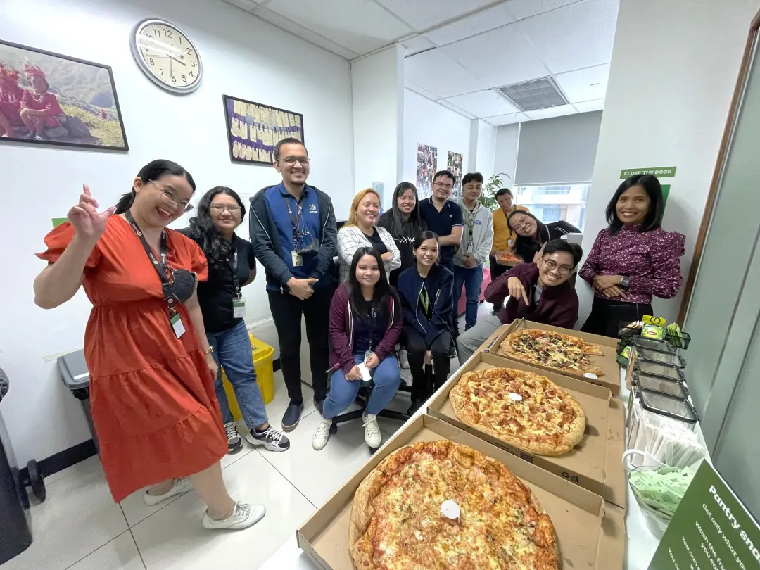 Group of coworkers gathered around a table with three large pizzas in an office room, celebrating Pizza FriYAY.