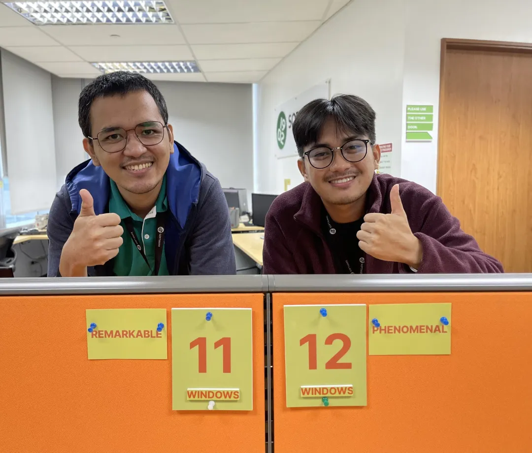 Two people behind orange cubicle panels labeled "11 WINDOWS REMARKABLE" and "12 WINDOWS PHENOMENAL" giving thumbs up