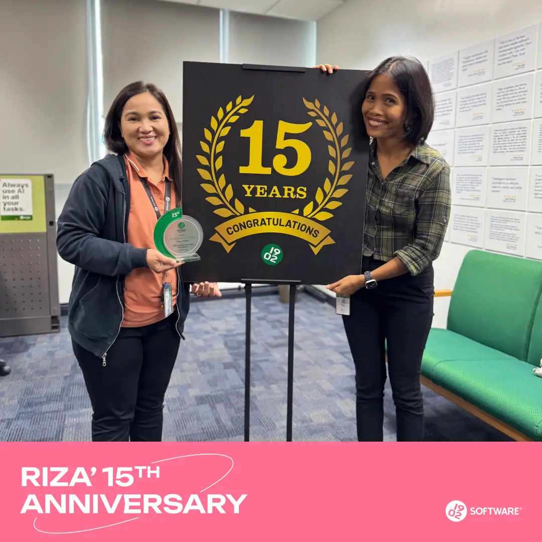 Two women holding a black sign with gold laurel wreath and text "15 Years Congratulations" celebrating Riza's 15th anniversary at 1902 Software.