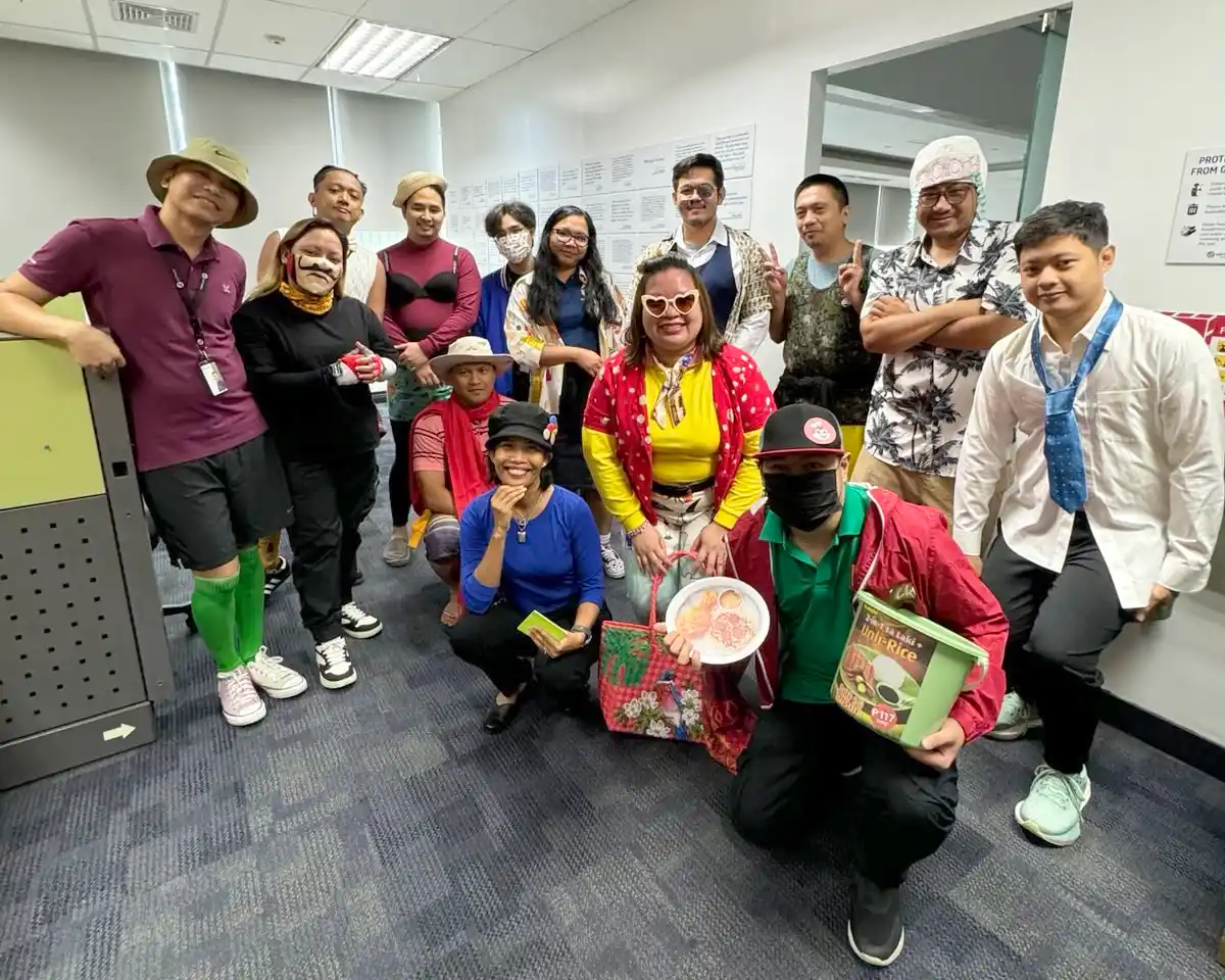Group of 14 coworkers in colorful casual and costume attire posing in an office hallway, some holding food containers.