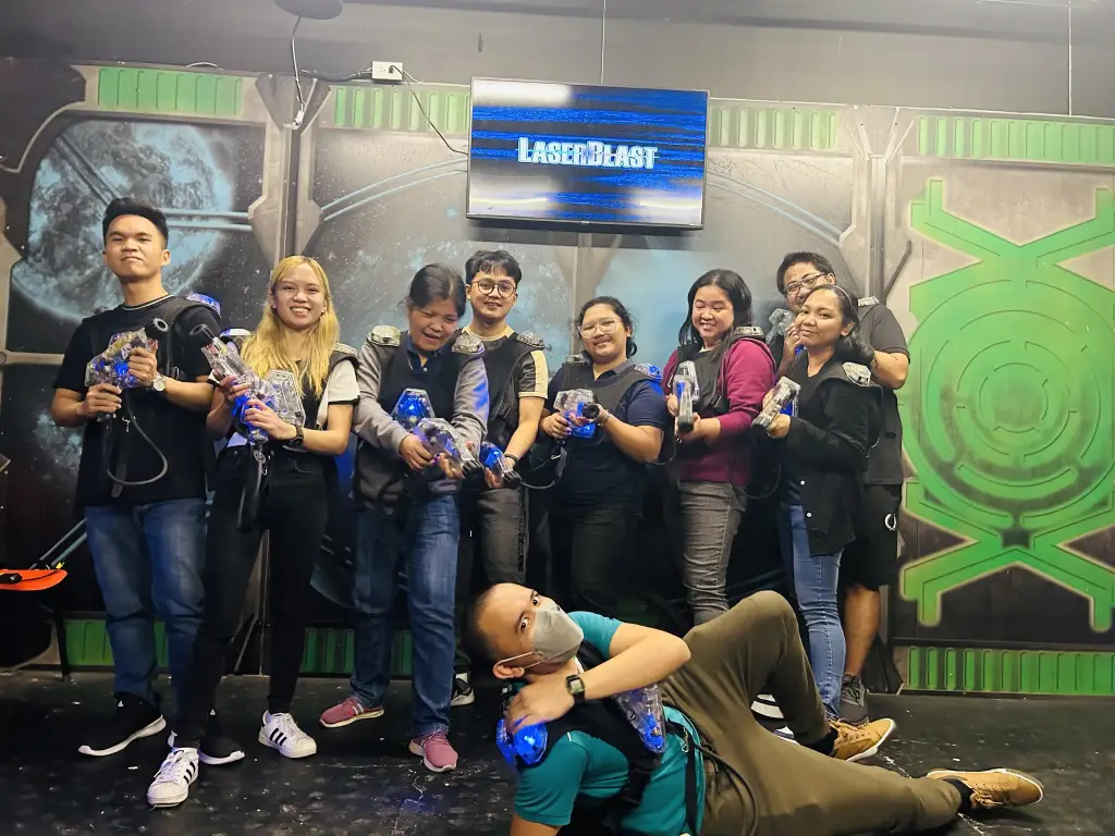 Group of coworkers wearing laser tag vests and holding laser guns posing in front of a LaserBlast sign during team building event