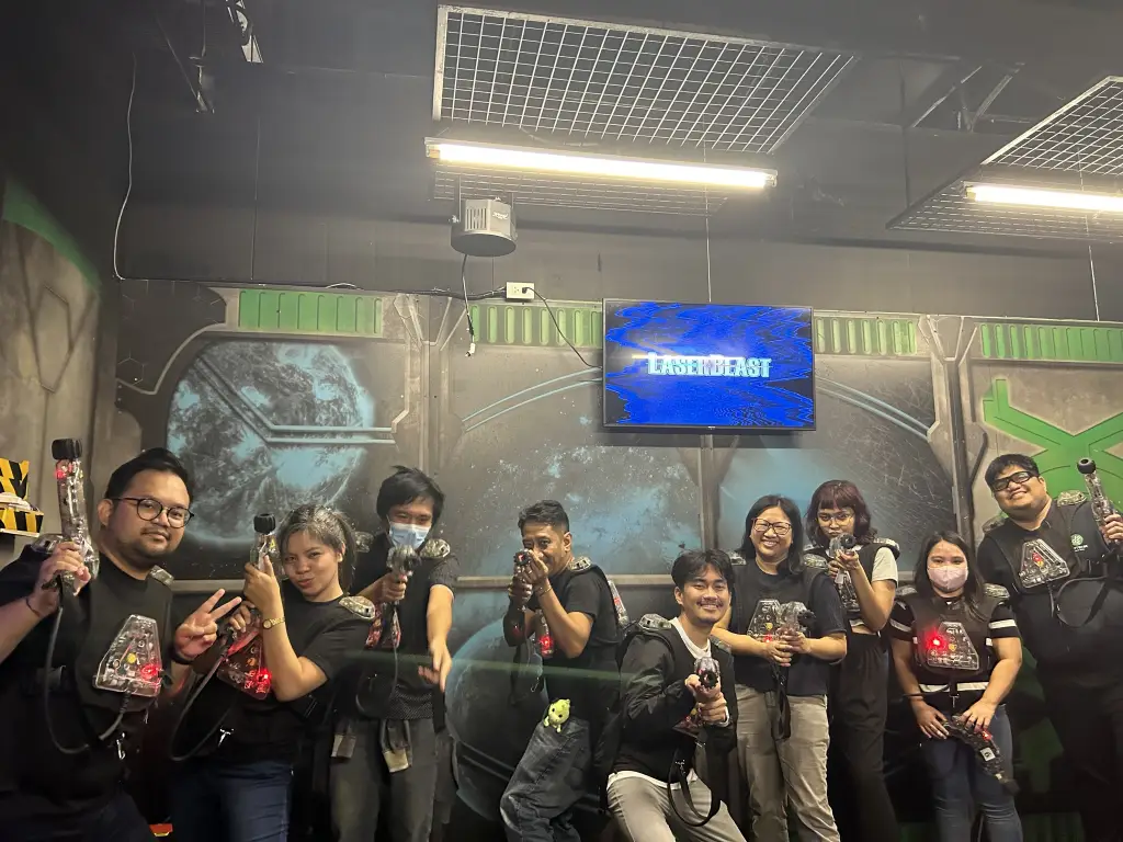 Group of people wearing laser tag vests and holding laser guns posing in front of a space-themed wall with a screen displaying "LASERBEAST"