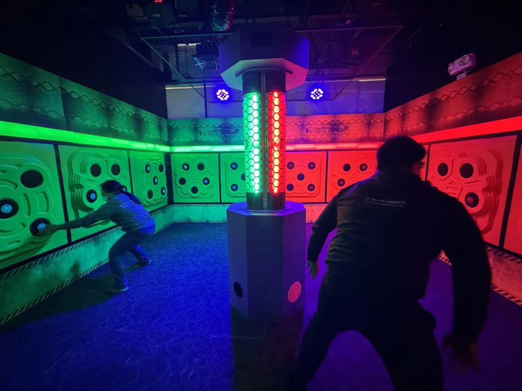 Two people playing an interactive light game in a neon-lit room with green and red panels and a central column with glowing lights.