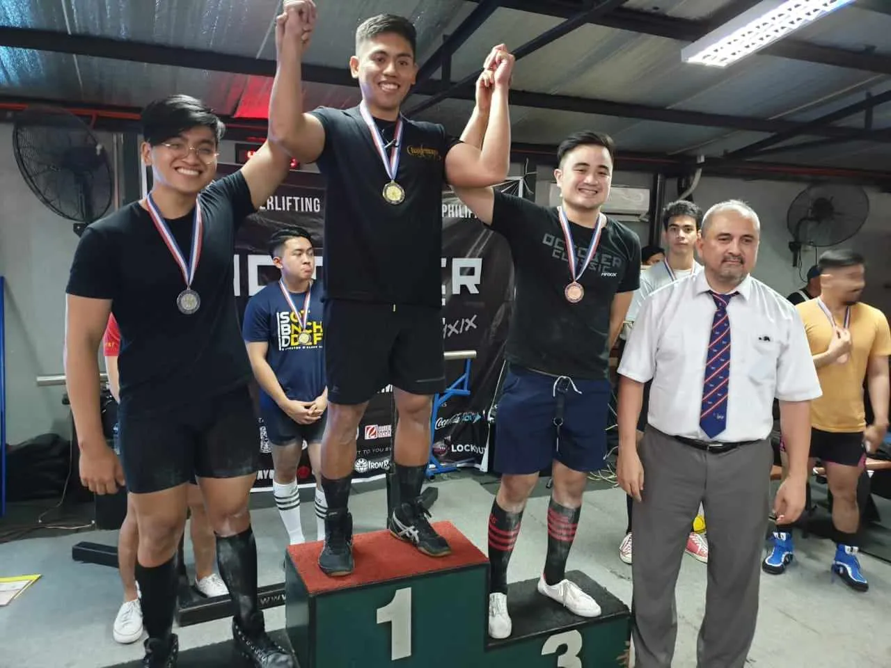 Three male athletes wearing medals stand on a winners' podium with a man in a white shirt and tie beside them in an indoor gym.