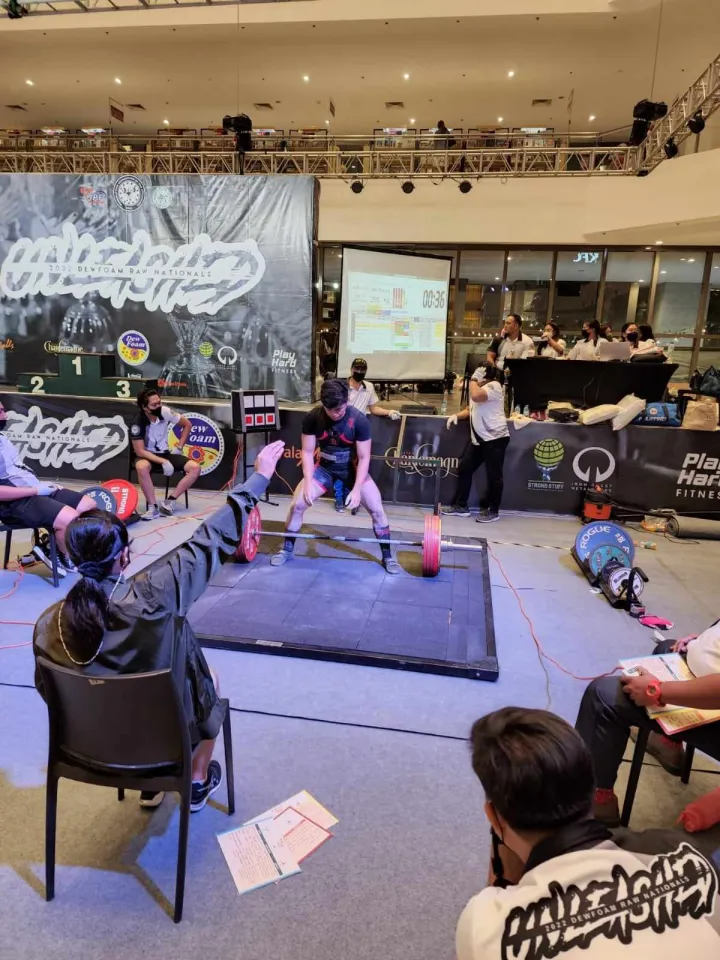 Weightlifting competition with athlete preparing to lift barbell, judges seated around, and scoreboard in background.