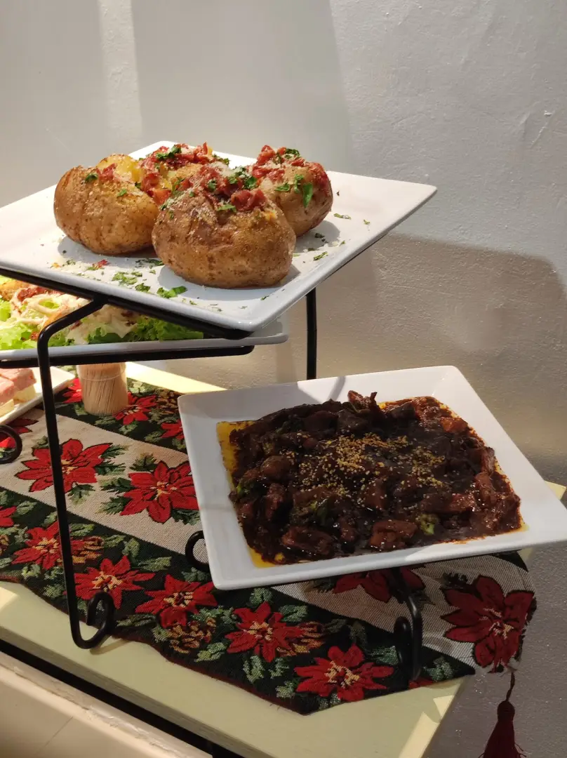 Four baked potatoes topped with bacon and herbs on a white plate above a plate of beef and broccoli with sesame seeds on a floral table runner