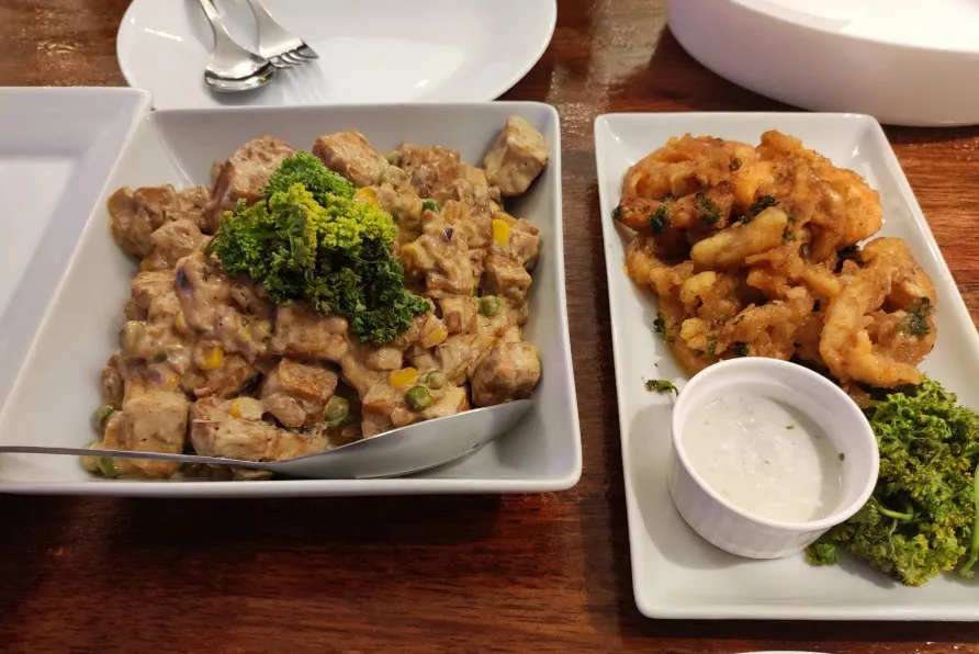Creamy tofu with mixed vegetables garnished with parsley in a square dish, breaded fried squid with dipping sauce and parsley on a rectangular plate.
