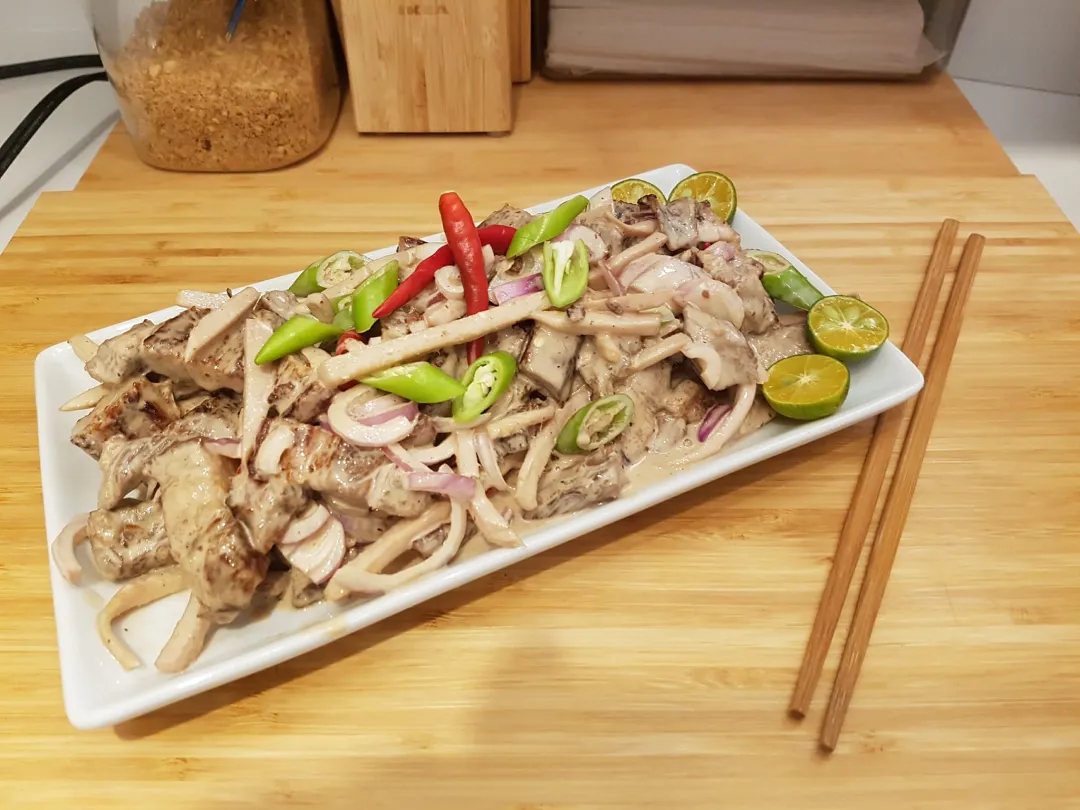 Filipino dish Dinakdakan with grilled pork, onions, chili peppers, and calamansi on a white rectangular plate with chopsticks on a wooden surface.