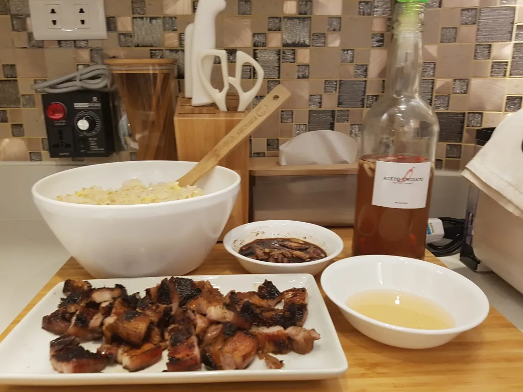 Grilled liempo slices on a white plate, garlic fried rice in a white bowl, dipping sauces, and a bottle of vinegar on a kitchen counter.