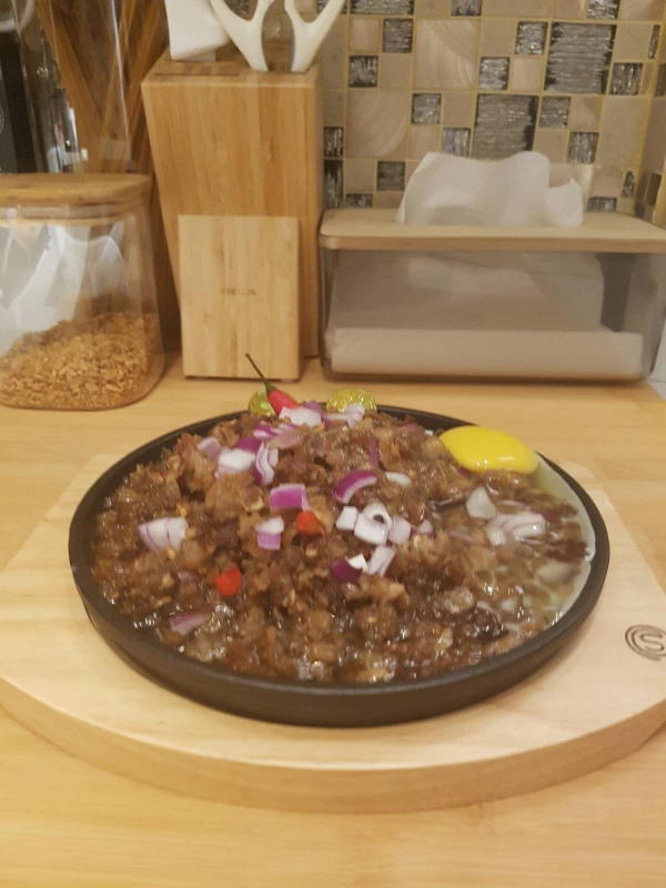Sizzling sisig dish topped with chopped red onions, chili, and a raw egg yolk on a wooden board in a kitchen setting