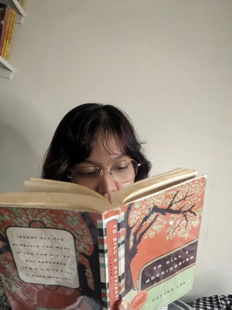 Person reading "To Kill a Mockingbird" by Harper Lee with the book cover visible showing a tree illustration and text.