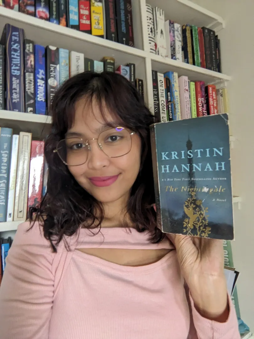 Person in pink top holding the book "The Nightingale" by Kristin Hannah in front of a bookshelf filled with books