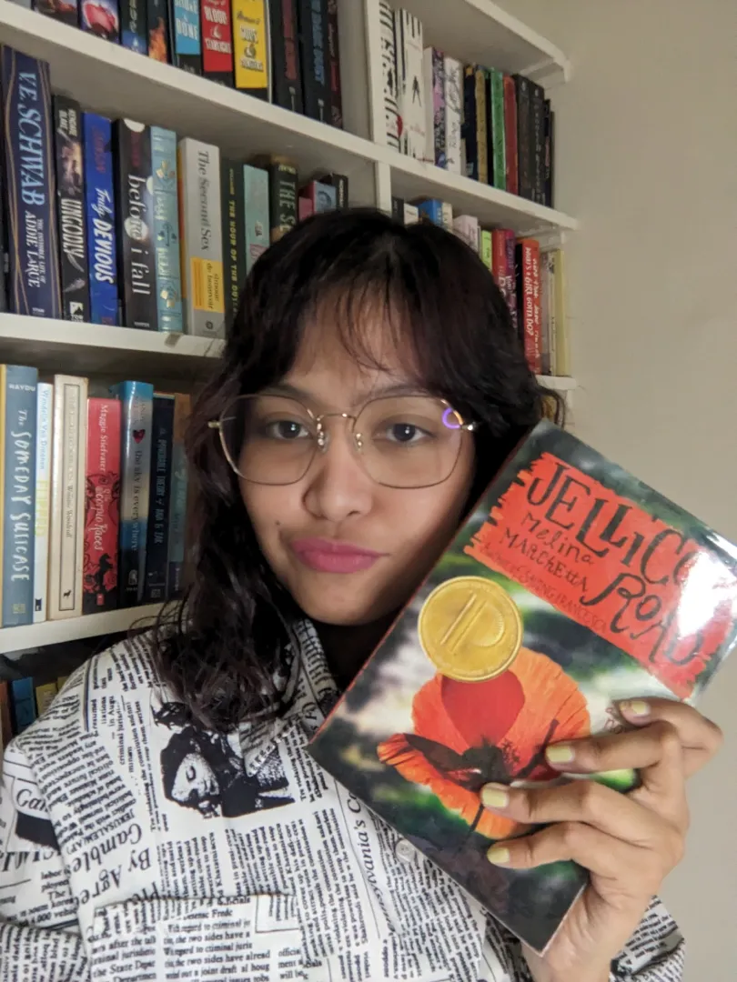 Person holding the book "Jellicoe Road" by Melina Marchetta in front of a bookshelf filled with various books