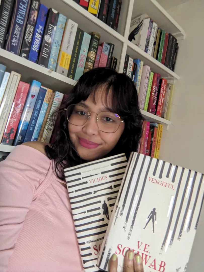 Person in pink top holding books "Vicious" and "Vengeful" by V.E. Schwab in front of a filled bookshelf