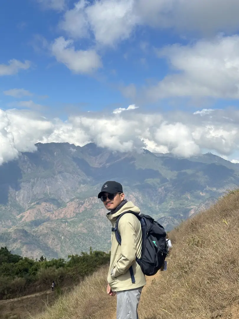 Person wearing a beige jacket and black cap hiking on a grassy slope with mountains and clouds in the background