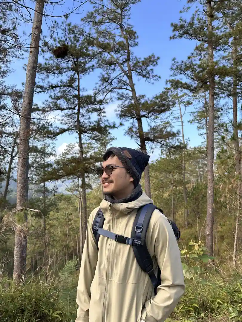 Person wearing a beige jacket, black beanie, and backpack standing in a forest with tall pine trees under a blue sky