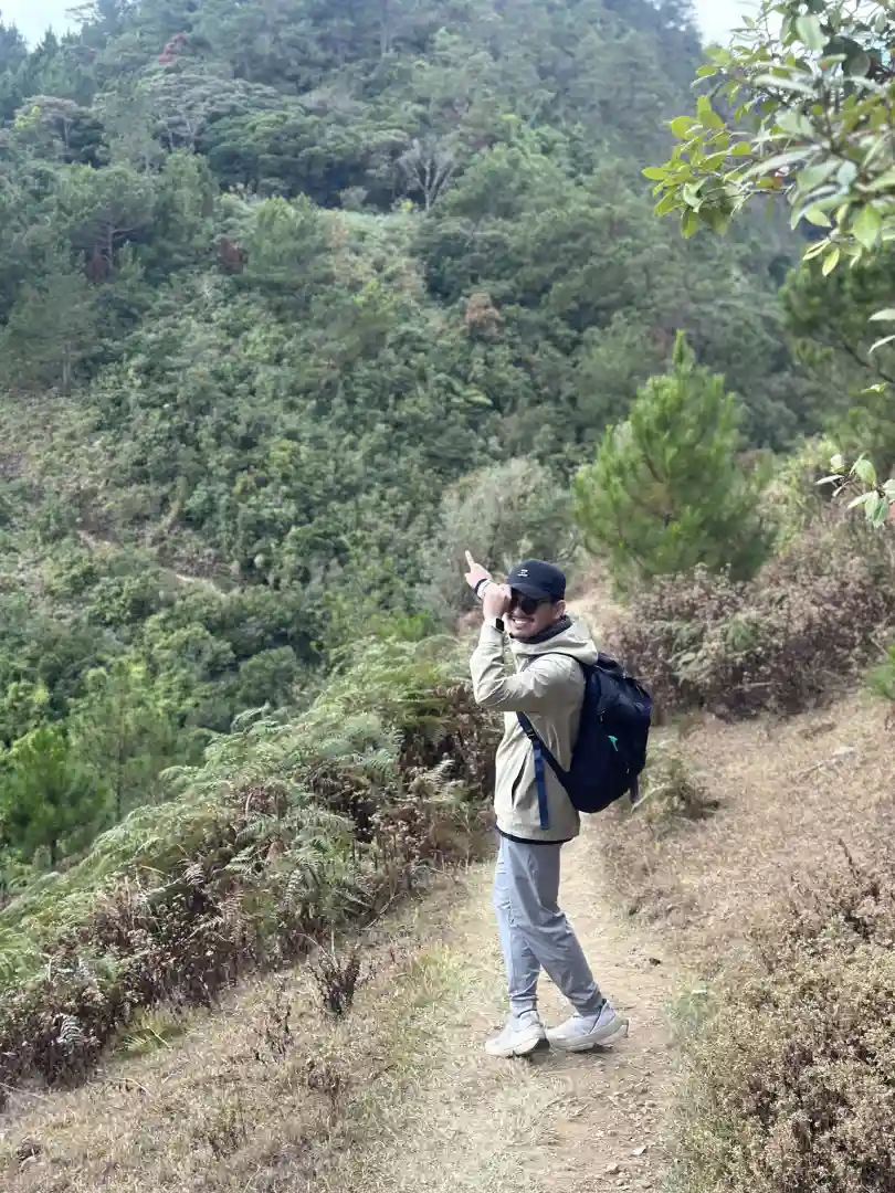 Person wearing a beige jacket and black cap pointing towards a green forested hillside while standing on a dirt trail