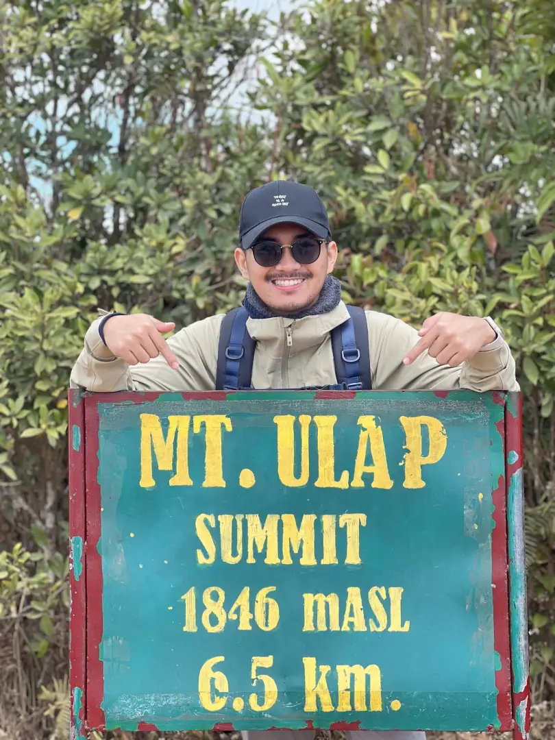Person wearing a cap and jacket pointing to a green and red sign reading "MT. ULAP SUMMIT 1846 MASL 6.5 km" with foliage background