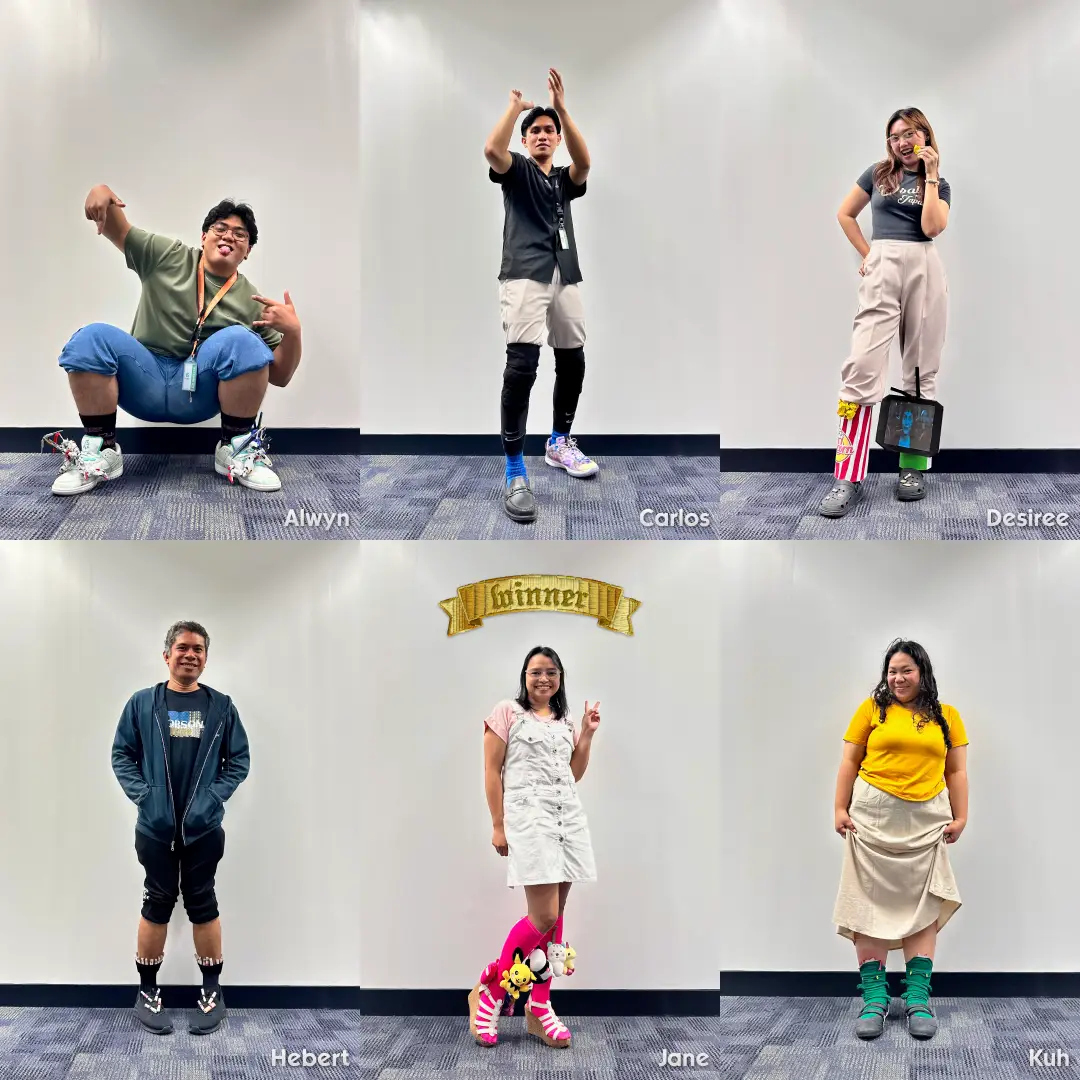 Six team members posing against a white wall with names below: Alwyn crouching, Carlos clapping, Desiree with popcorn and screen on leg, Hebert standing, Jane with pink socks and "winner" banner above, Kuh standing in yellow shirt.
