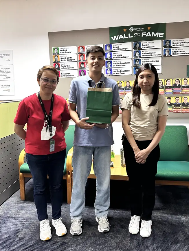 Three people standing in front of the 1902 Software Wall of Fame: a female in red on the left, Adrian in the center holding a green gift bag, and a female on the right