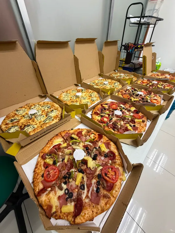 A long table in an office setting filled with several open boxes of Angel's Pizza, featuring various toppings like spinach and cheese, and meat lovers with tomatoes and olives.