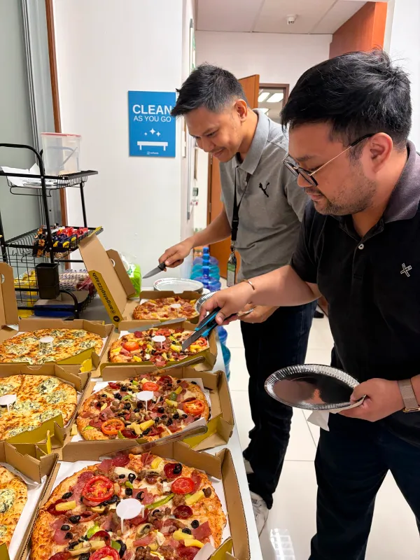 Two office employees smiling as they serve themselves slices of pizza from several open boxes on a table.
