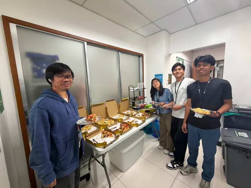 Four interns posing and smiling for the camera while holding plates with pizza slices in an office pantry area.