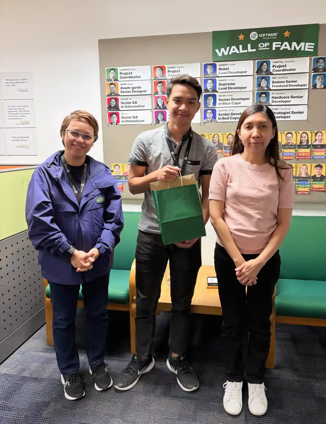 Three employees standing in an office in front of a wall of fame display, with one holding a green gift bag and chairs behind them, showcasing a workplace recognition moment.