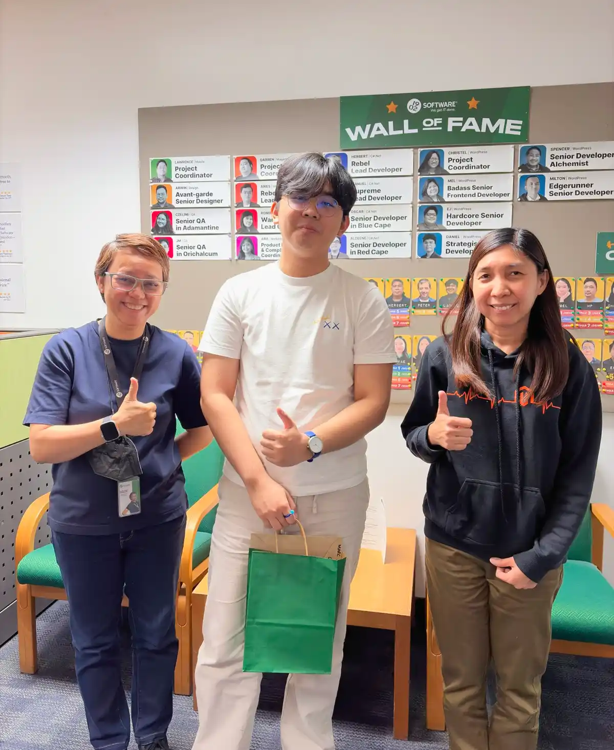 Three people posing in an office area in front of a "Wall of Fame" board with staff profiles, each giving a thumbs-up gesture, while the person in the center holds a green paper gift bag