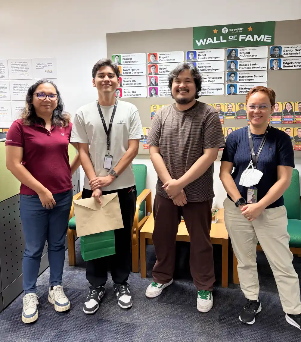 Four people standing together in an office in front of a company Wall of Fame board, posing during an intern recognition or workplace achievement moment, with one holding an envelope and chairs visible in the background.